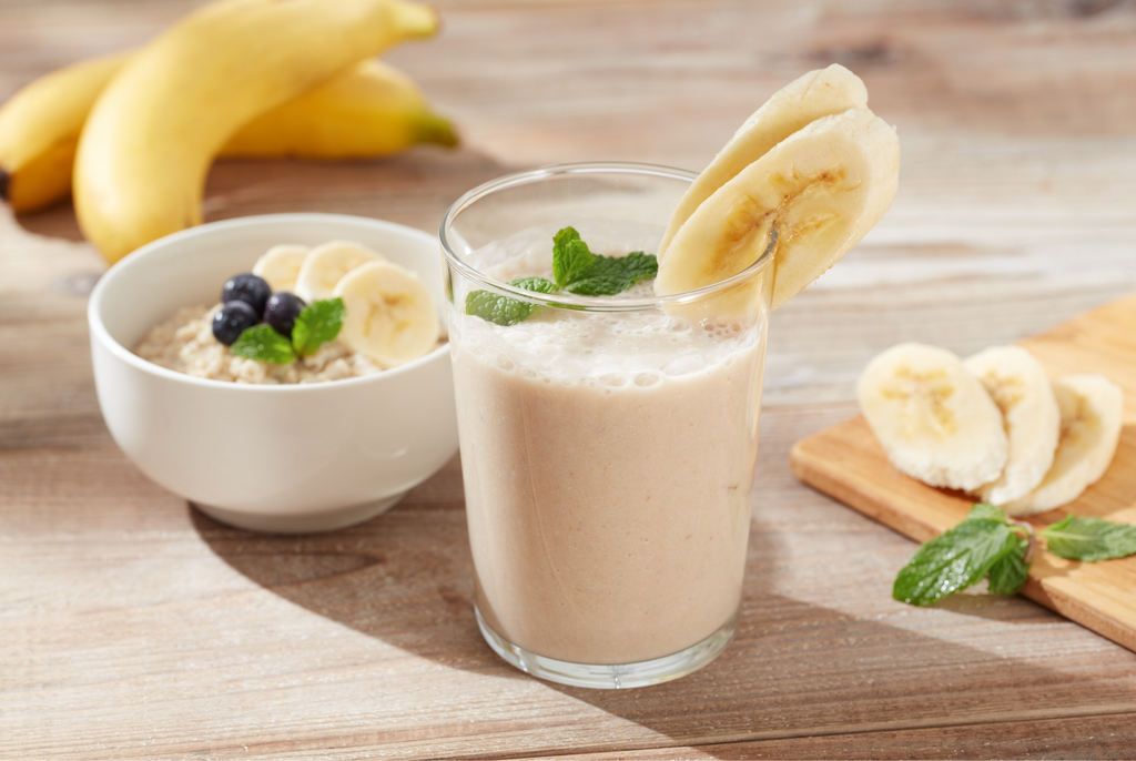 Glass of banana smoothie with a bowl of oatmeal and bananas on a wooden table.