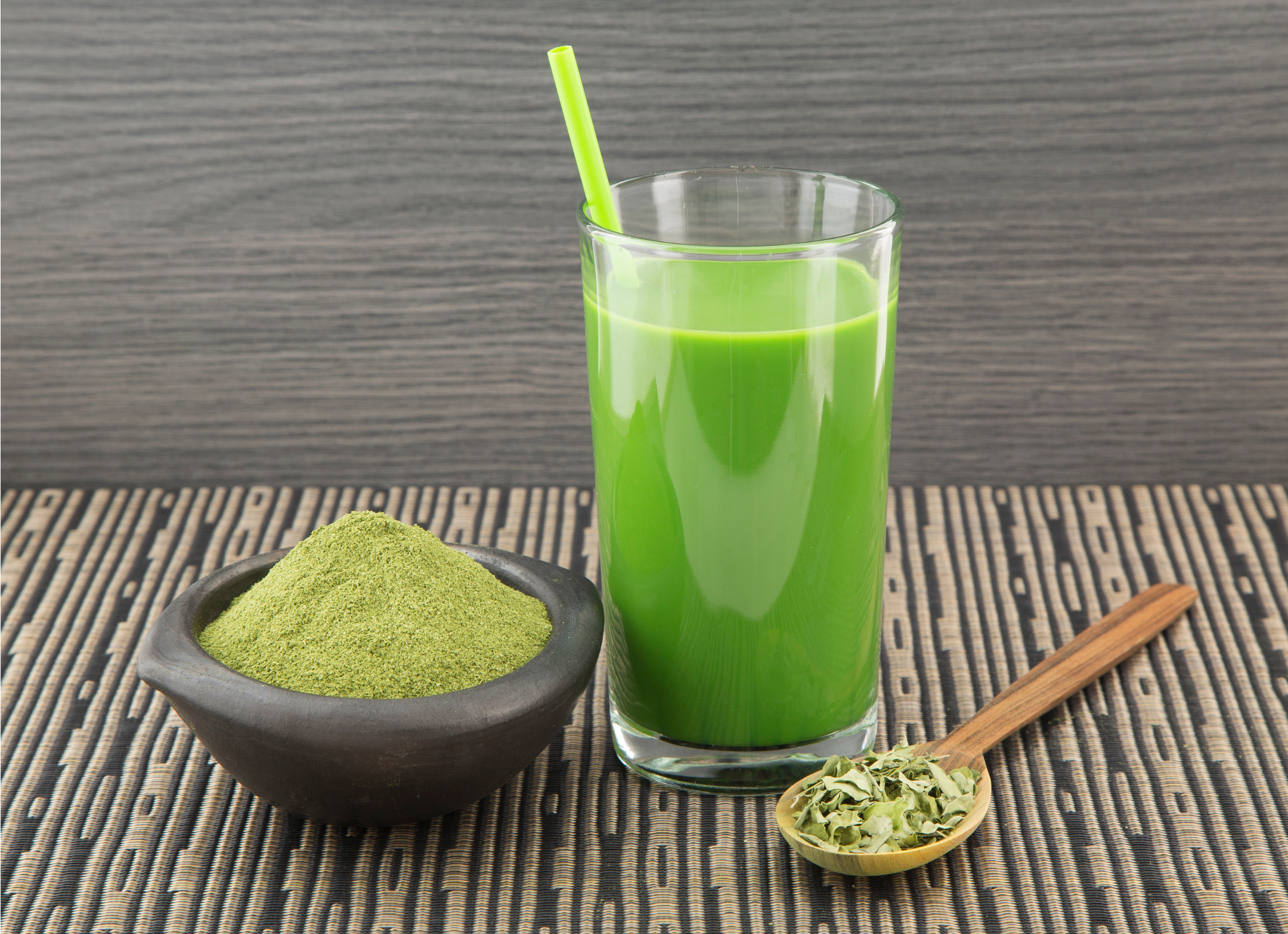 Green Moringa smoothie in a glass with a bowl of green moringa powder and a wooden spoon on a textured surface.
