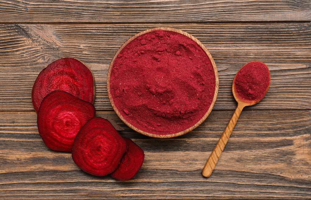 Red beetroot powder in a bowl with sliced beets and a wooden spoon on a wooden surface.